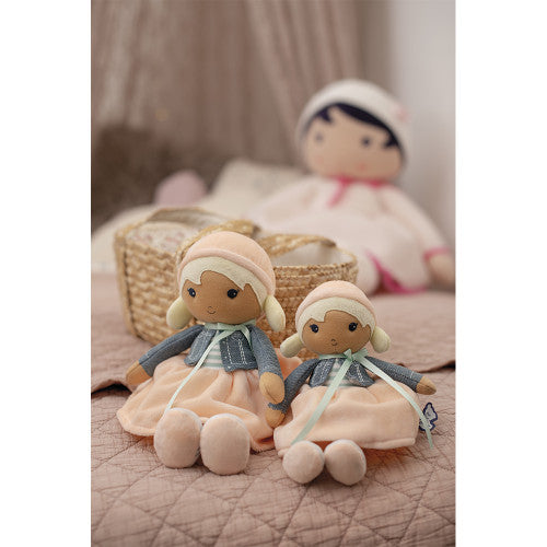 Two dolls sitting on a bed, with a basket and another doll in the background.